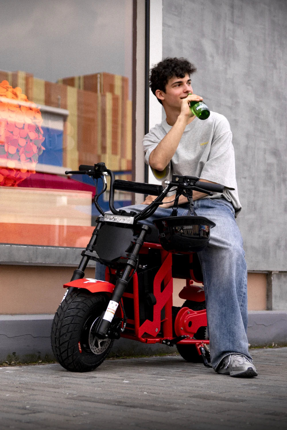Young man relaxing on a red FRP M3 electric mini bike by a shop window